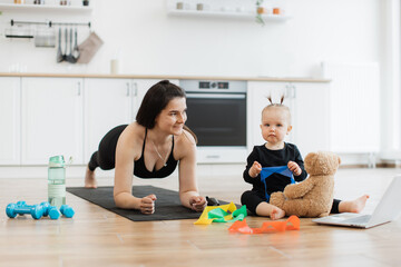 Athletic woman holding plank pose on mat while sweet kid choosing color of resistance band for teddy bear. Sporty mother and daughter in yoga outfit getting bonding experience in home interior.