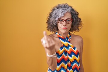 Middle age woman with grey hair standing over yellow background showing middle finger, impolite and...