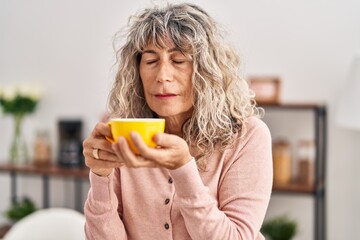 Middle age woman smelling cup of coffee sitting on table at home