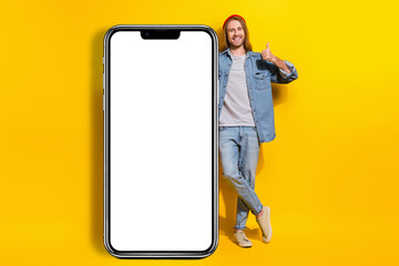 Full length photo of positive guy dressed jeans shirt trousers lean on smart phone showing thumb up isolated on yellow color background