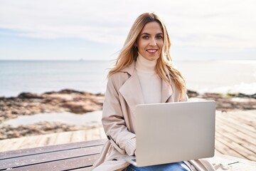 Young blonde woman using laptop sitting on bench at seaside