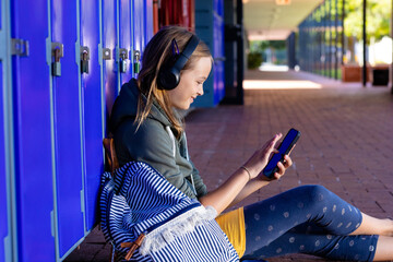 Smiling caucasian schoolgirl in headphones using smartphone sitting by lockers at school