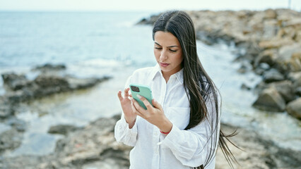 Young beautiful hispanic woman using smartphone with serious expression at seaside