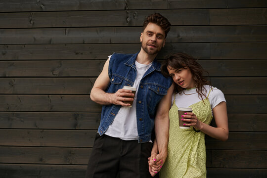 Trendy brunette couple in summer outfits holding hands and takeaway coffee and looking at camera while standing near wooden house in rural setting, carefree moments concept - Powered by Adobe