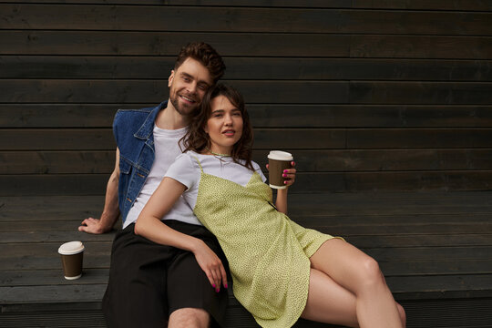 Fashionable stylish and romantic couple in summer outfits smiling at camera while holding coffee to go and sitting near wooden house in rural setting, carefree moments concept, tranquility - Powered by Adobe