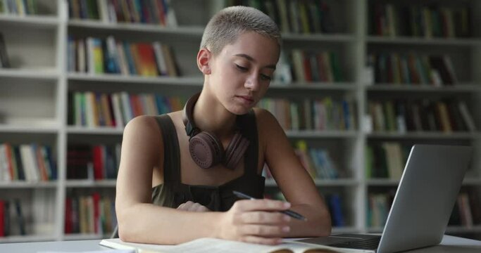 Serious thoughtful trendy looking student girl studying in university library, sitting at laptop, writing notes, using app, turning look away, thinking, catching exciting creative idea