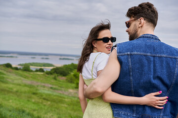 Cheerful and fashionable brunette woman in sunglasses and sundress hugging bearded boyfriend in denim vest while standing on blurred grassy hill at background, countryside retreat concept