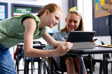 Happy caucasian schoolgirl in wheelchair with her friend using tablet in school classroom