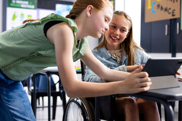 Happy caucasian schoolgirl in wheelchair with her friend using tablet in school classroom