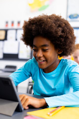 Happy african american schoolboy sitting at desk and using tablet in school classroom