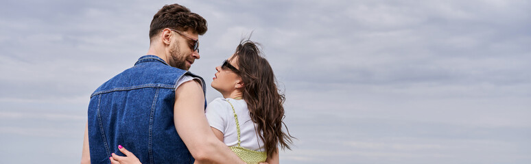 Side view of trendy romantic couple in sunglasses and summer outfits hugging and looking at each other while standing with cloudy sky at background, countryside leisurely stroll, banner