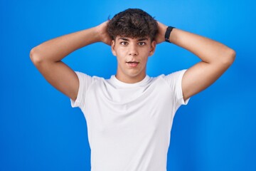 Hispanic teenager standing over blue background crazy and scared with hands on head, afraid and...