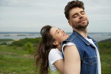 Fototapeta premium Cheerful brunette woman with closed eyes standing next to stylish bearded boyfriend in denim vest and spending time together with landscape at background, love story and countryside adventure