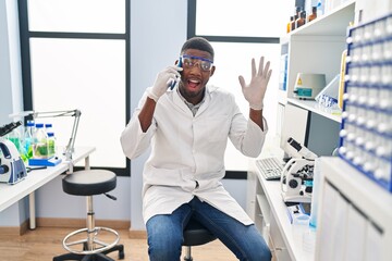 African american man working at scientist laboratory speaking on the phone scared and amazed with open mouth for surprise, disbelief face