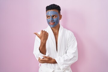 Young hispanic man wearing beauty face mask and bath robe smiling with happy face looking and...