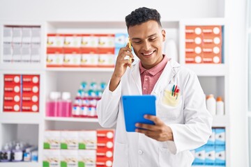 Young latin man pharmacist talking on smartphone holding pills bottle at pharmacy