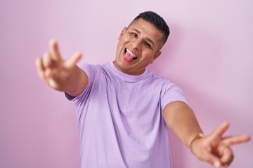 Young hispanic man standing over pink background smiling with tongue out showing fingers of both hands doing victory sign. number two.