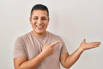 Hispanic young man standing over white background amazed and smiling to the camera while presenting with hand and pointing with finger.