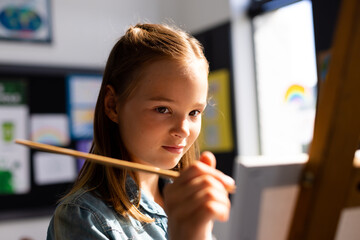 Fototapeta premium Happy caucasian schoolgirl painting using brush and easel in school art class