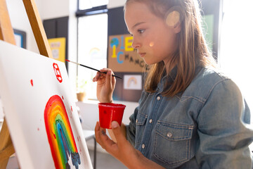 Fototapeta premium Caucasian schoolgirl painting using brush and easel in school art class