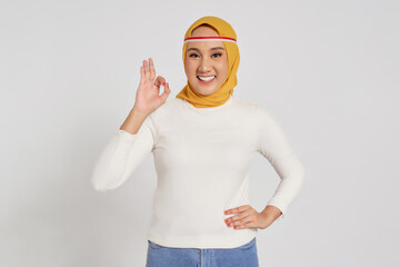 Smiling happy young Asian Muslim woman wearing a hijab showing ok sign isolated over white...