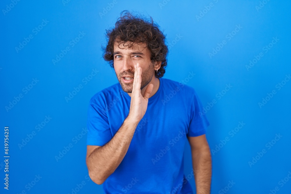 Canvas Prints Hispanic young man standing over blue background hand on mouth telling secret rumor, whispering malicious talk conversation