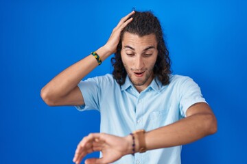 Young hispanic man standing over blue background looking at the watch time worried, afraid of getting late