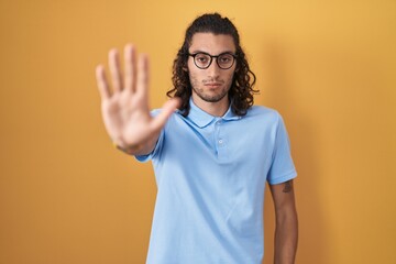 Young hispanic man standing over yellow background doing stop sing with palm of the hand. warning...