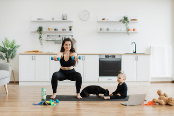 Attractive young mom practising squats with dumbbells while calm baby girl having fun with small...