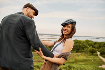 Cheerful brunette woman in vest and newsboy cap touching bearded boyfriend in jacket and looking away with nature and overcast sky at background, fashion-forwards in countryside