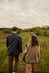 Brunette and fashionable woman in vest and newsboy cap holding hand of boyfriend in jacket while walking together on meadow with nature and sky at background, fashion-forwards in countryside
