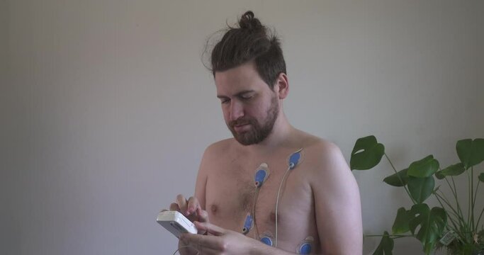Man with ECG/EKG leads on his chest looks down at heart monitoring device in clinic or home setting