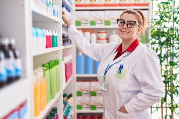 Young beautiful plus size woman pharmacist smiling confident holding product on shelving at pharmacy