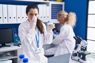 Young hispanic woman working at scientist laboratory covering mouth with hand, shocked and afraid...