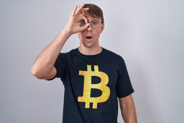 Caucasian blond man wearing bitcoin t shirt doing ok gesture shocked with surprised face, eye looking through fingers. unbelieving expression.