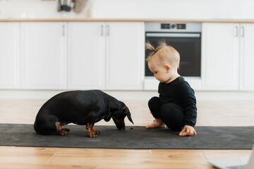 Adorable little girl in black outfit crouching near funny dog on yoga mat in modern kitchen. Sweet...