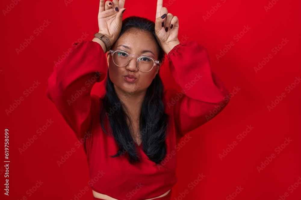 Poster asian young woman standing over red background doing funny gesture with finger over head as bull hor