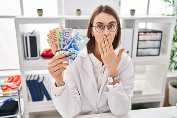 Young caucasian doctor woman holding canadian dollars banknotes covering mouth with hand, shocked...