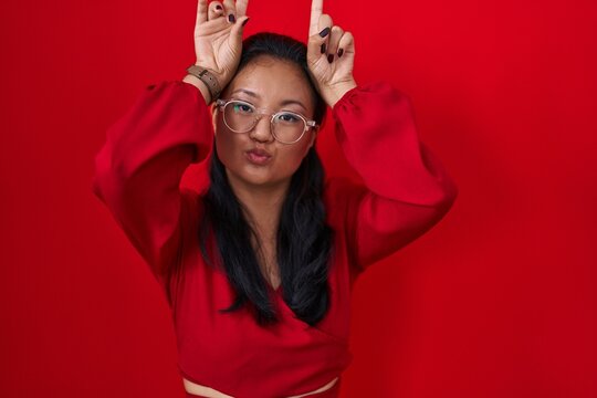 Asian young woman standing over red background doing funny gesture with finger over head as bull horns