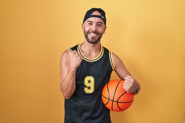 Middle age bald man holding basketball ball over yellow background very happy and excited doing...