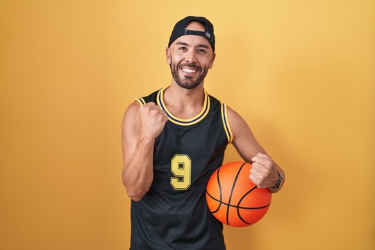 Middle age bald man holding basketball ball over yellow background very happy and excited doing winner gesture with arms raised, smiling and screaming for success. celebration concept.