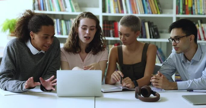 Class project leader presenting study research content on laptop to classmates. Diverse group of college students sitting at computer in library, talking, discussing homework tasks