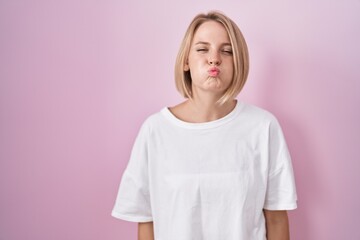 Young caucasian woman standing over pink background puffing cheeks with funny face. mouth inflated with air, crazy expression. - obrazy, fototapety, plakaty