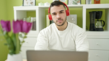 Young hispanic man using laptop and headphones sitting on table at dinning room