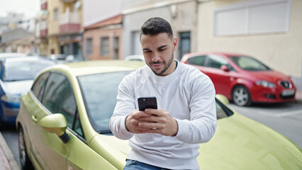 Young hispanic man using smartphone sitting on car at street