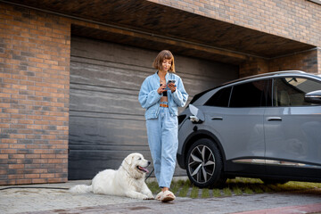 Young woman stands with her dog and using smart phone while charging her electric car near garage of her house. Concept of modern lifestyle and sustainability - 619762637