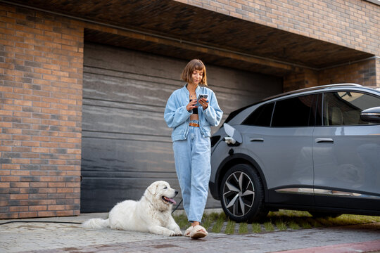 Young woman stands with her dog and using smart phone while charging her electric car near garage of her house. Concept of modern lifestyle and sustainability