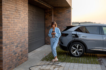Young woman using smart phone while charging her electric car near garage of her house. Concept of...