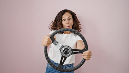 Middle age hispanic woman smiling confident using steering wheel as a driver over isolated pink background