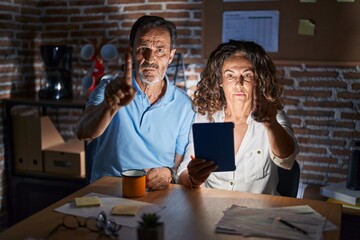 Middle age hispanic couple using touchpad sitting on the table at night pointing with finger up and angry expression, showing no gesture